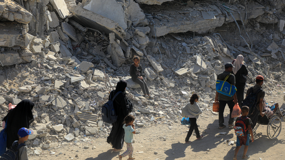  A group of people walk past rubble carrying sparse belongings. A young girl sits on the rubble watching them pass