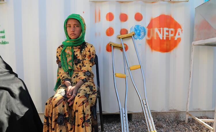 A girl in a yellow dress and green headscarf sits on a chair outside a metal wall bearing the UNFPA logo. Beside her are two crutches.