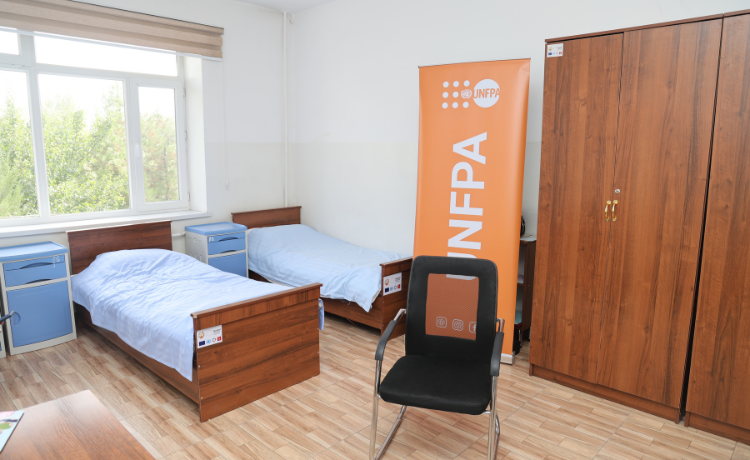 A room is made up of two single beds, bedside lockers, a wardrobe, a chair and a UNFPA banner. Behind the beds there is a window with a view of some trees.