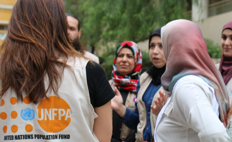 A woman wearing a vest with a UNFPA logo speaks to a group of women and one man in an outdoor courtyard.