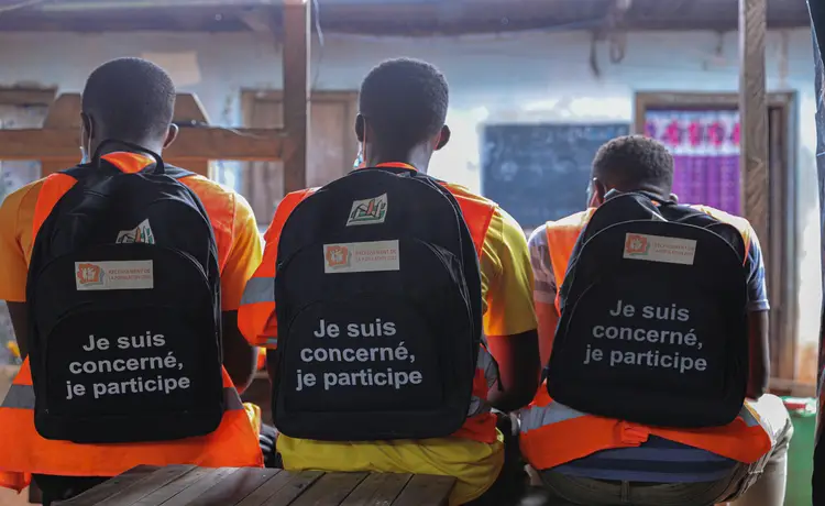 A group of males sit on a bench with backpacks on.