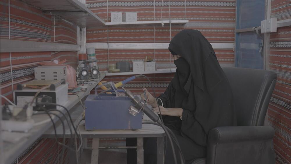 A woman in a black niqab and abaya sits at a workdesk, holding tools