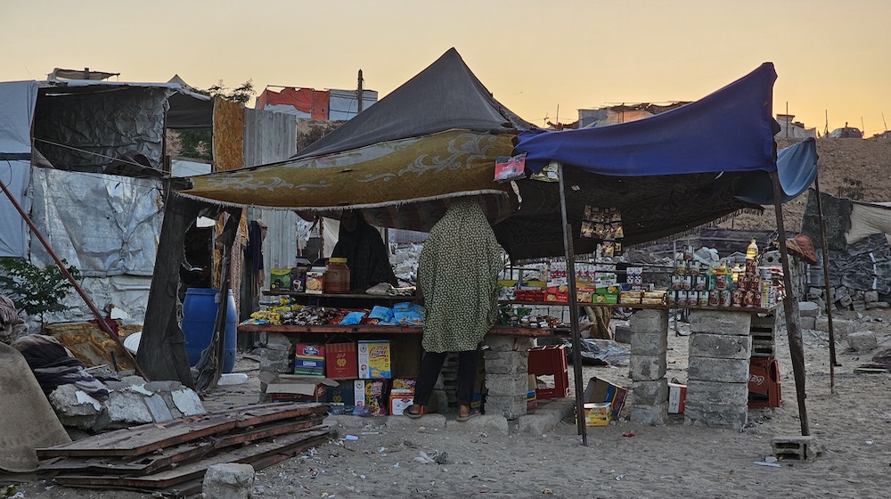 Des stocks de nourriture emballée sont empilés sur des tables de fortune sous des tentes dans un camp informel. On distingue une femme de dos qui porte un châle à motif.]