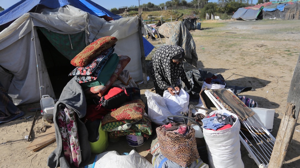 Une femme portant un foulard à motif est penchée sur des piles de sacs et d’affaires, devant une tente bleue et blanche plantée dans un champ sec et poussiéreux