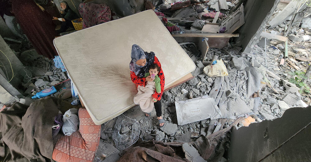 A woman cradles a child on her lap, sitting on a bare mattress surrounded by rubble and debris