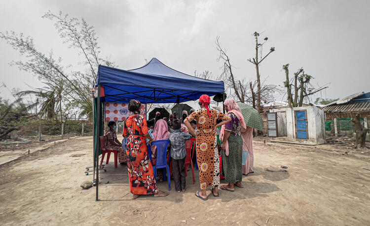 A group of women and one boy gather around a small blue tented table]