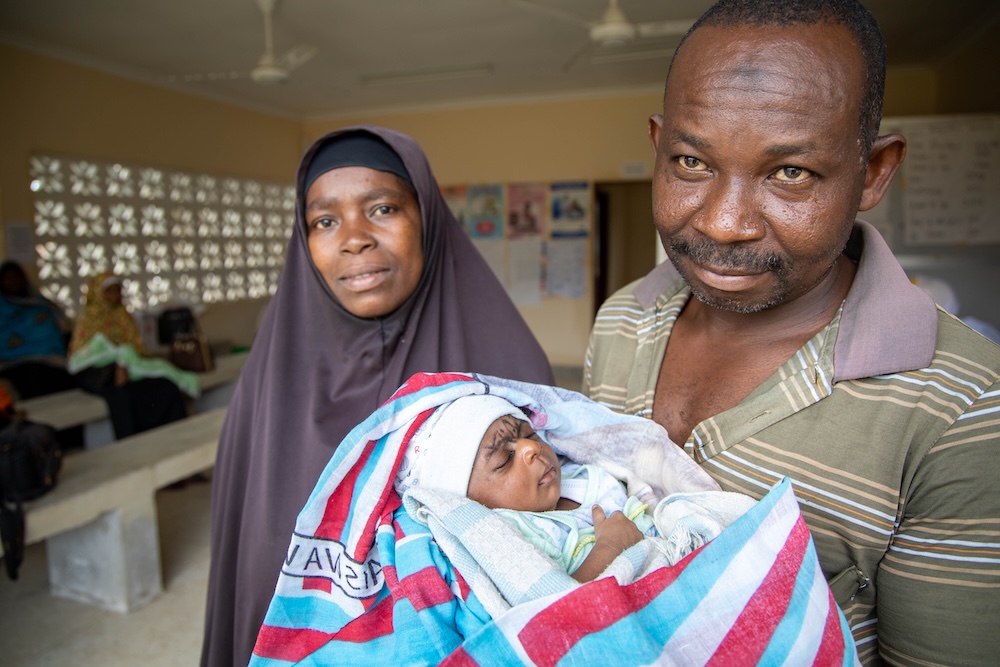 A man in a striped polo shirt holds his infant child, who is wrapped in a blanket. The child’s mother, wearing a black hijab, stands to the side smiling