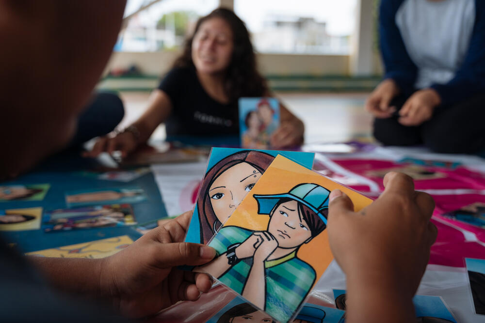 A group of people sit at a table and play a game with cards. The cards show drawings of people showing different emotions.