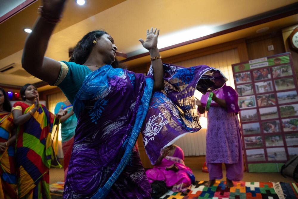 Maimouni, a member of Bangladesh’s hijra group, is dressed in pink and purple clothing and sits and speaks among a group.