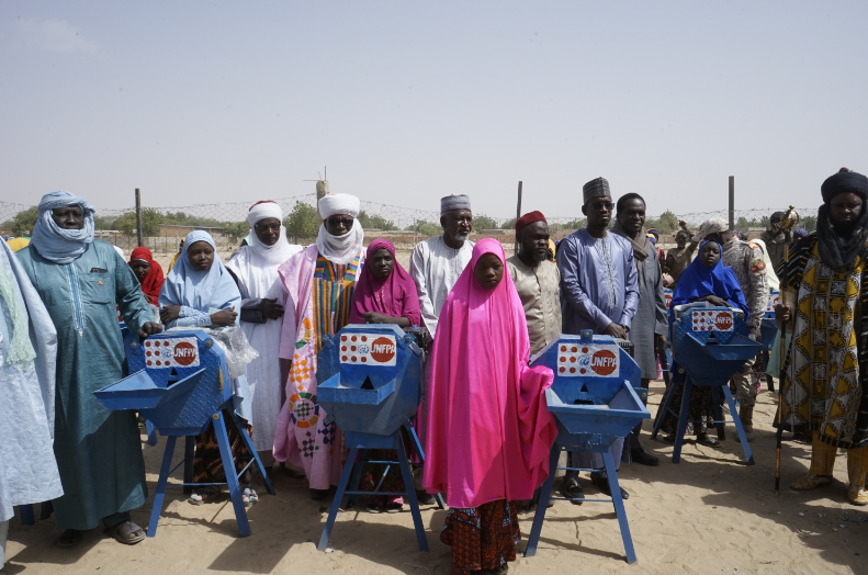 Teenage girls and village elders stand beside seed grinding mills with UNFPA logo