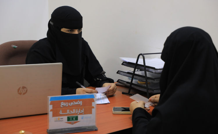 wo women in black niqabs speak across a desk with a laptop and filing tray on it]