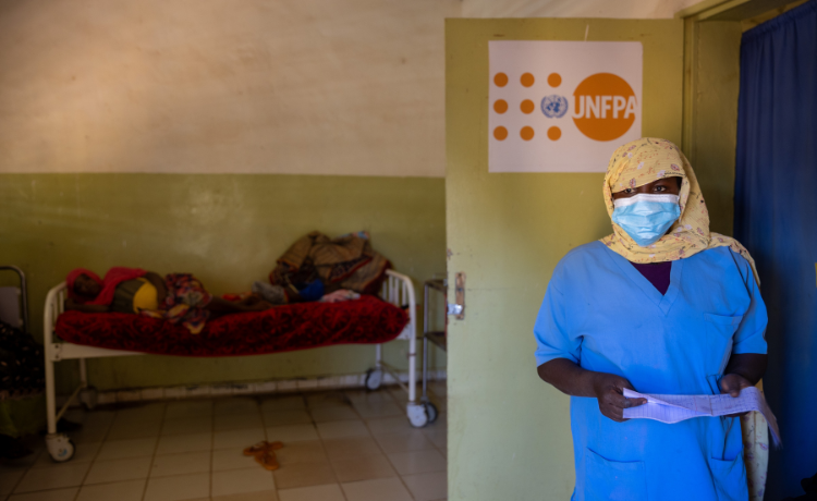 A female health worker in blue scrubs stands in the doorway of a room, where a pregnant woman is lying on a hospital bed