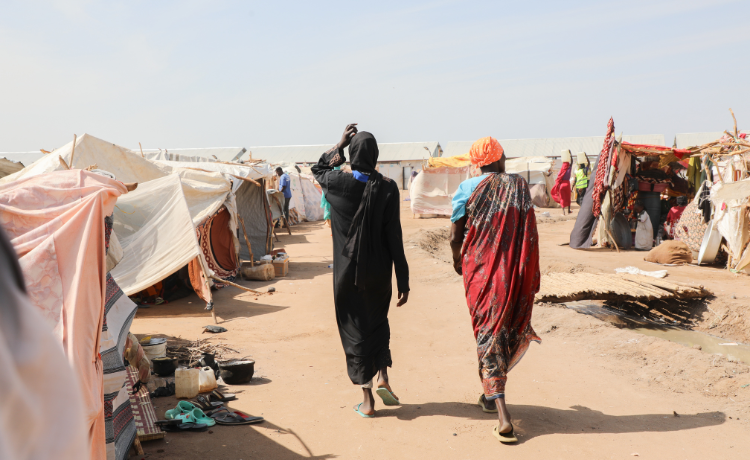 Two people seen from behind walk along a sandy path with makeshift tents on wither side