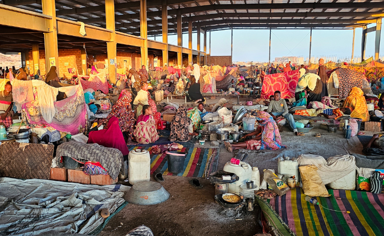 A view of a semi openair makeshift shelter, full of people, tents, bedding and belongings