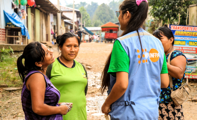 Four women chat in a circle. One of them has a UNFPA-branded blue vest. They are all standing in a village street