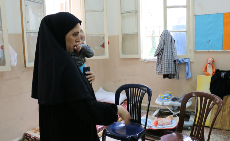 A woman in a black headscarf holds a baby in a classroom. A mattress and baby bed are on the floor.