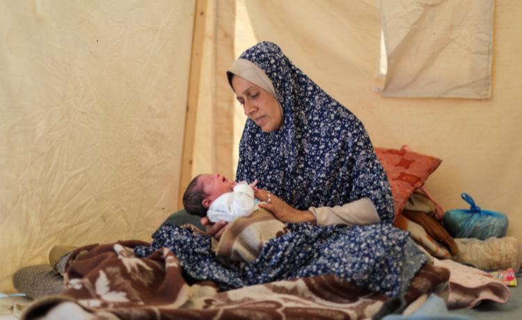 A woman in a patterned blue dress holds a crying newborn baby inside a tent.