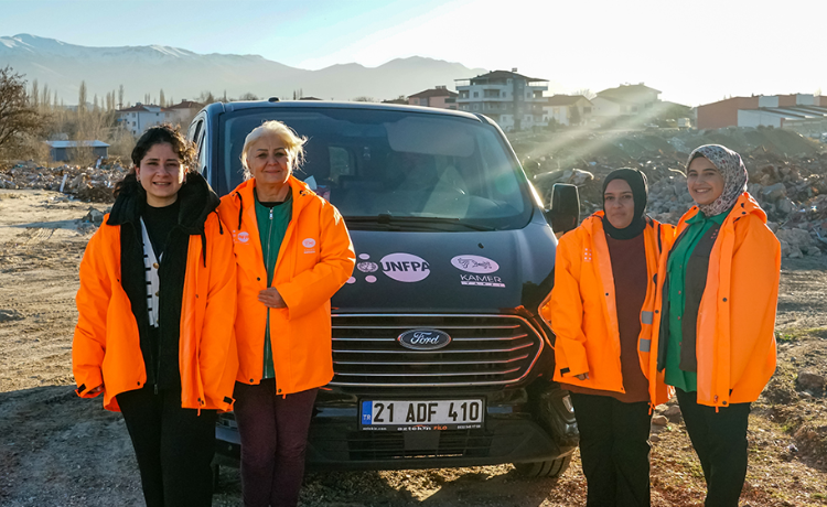 Quatre femmes en vestes orange fluo se tiennent devant une voiture, au milieu de débris provoqués par le sésisme de 2023 en Türkiye.