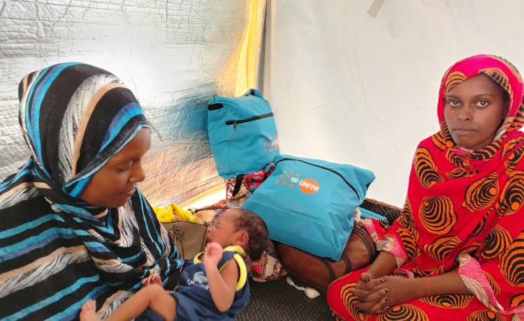 A woman dressed in red smiles lightly into the camera as she sits in a tent, with UNFPA bags visible behind her. Another woman in blue and black stripes holds a newborn crying baby.