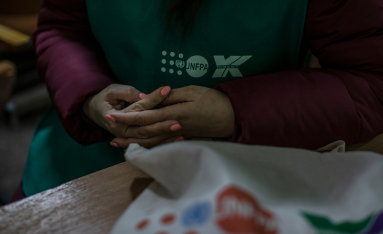 Una mujer que con esmalte de uñas naranja junta sus manos en su regazo al sentarse en un escritorio, con una pieza de vestir verde en que se aprecia el nombre del UNFPA