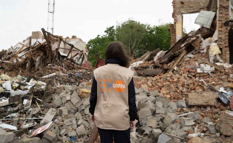 Una mujer de pie frente a casas convertidas en escombros da la espalda a la cámara con un chaleco del UNFPA