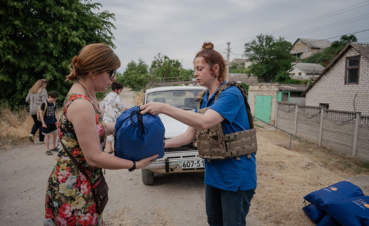 A woman in a combat vest hands a blue bag of UNFPA supplies to a woman in a flowery dress