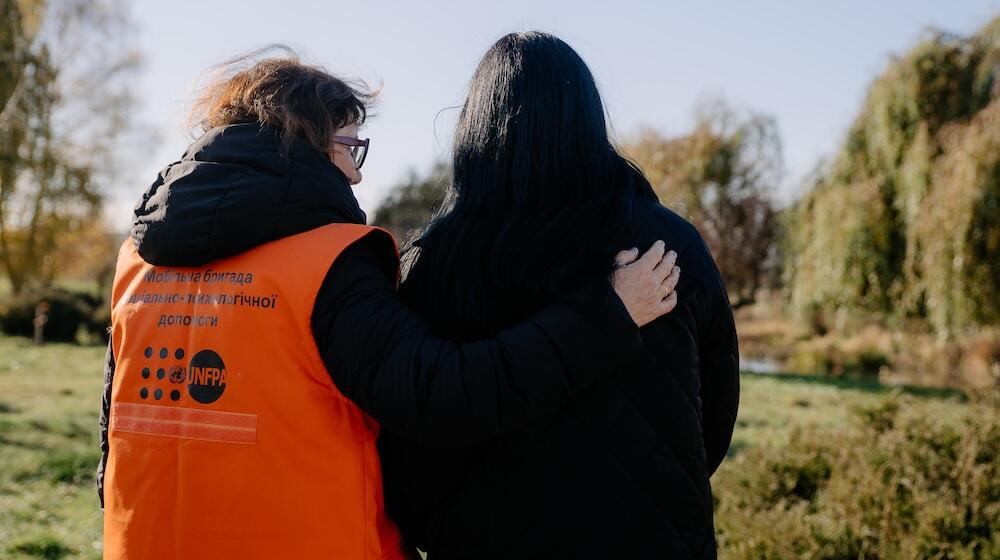 A woman wearing a UNFPA branded vest embraces another woman. 