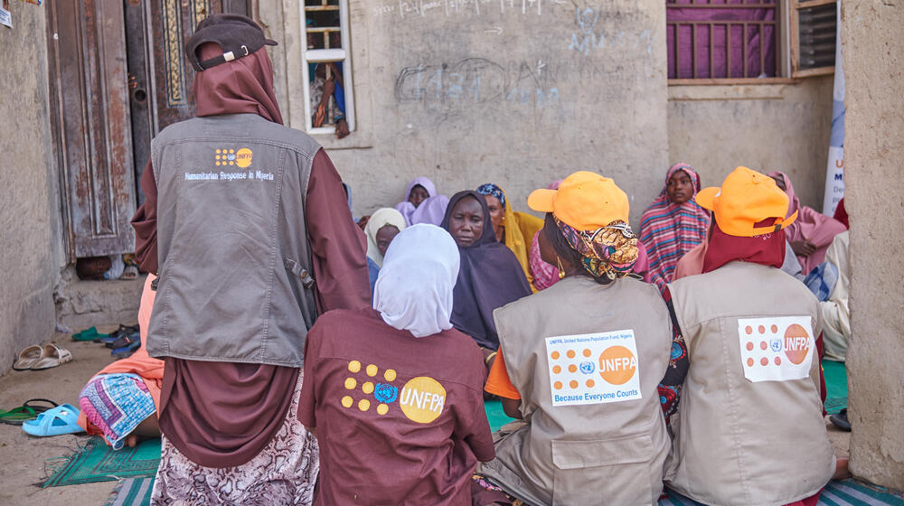 Des femmes portant des vêtements brodés du logo de l’UNFPA parlent à un groupe d’autres femmes assises sur des couvertures, dans une cour.