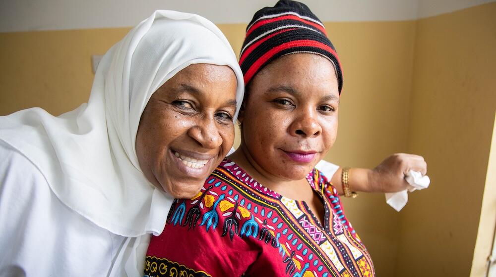 A woman in a white head scarf places her arm around a woman in a bright red and yellow dress. Both are smiling broadly.