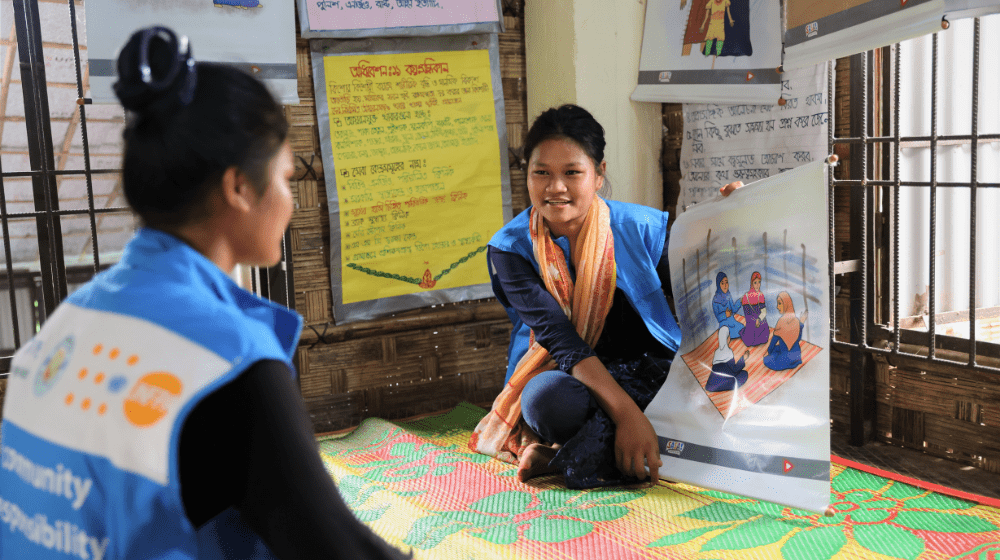  A smiling young woman holds a poster showing a group of women sitting together on a mat.