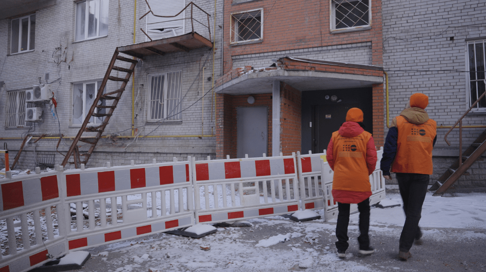 Two people in orange UNFPA-branded vests and hats walk towards a heavily damaged building