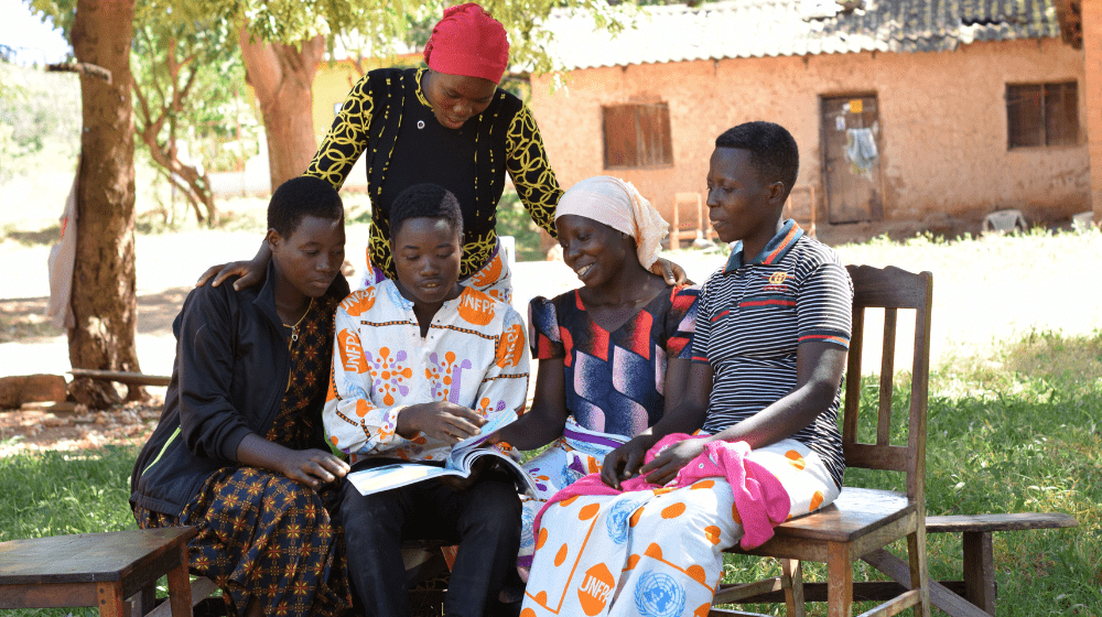  Young people crowd together in the shade of a courtyard to look at a book together. One wears a shirt with the UNFPA logo. Three others wear a wrap skirt with the UNFPA logo