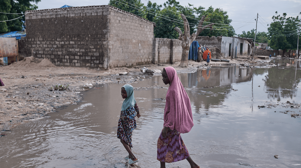 Un homme est assis sur un parpaing devant les décombres causés par les inondations, des tissus sont suspendus sur un fil à linge. Il porte un t-shirt gris floqué du logo UNFPA.