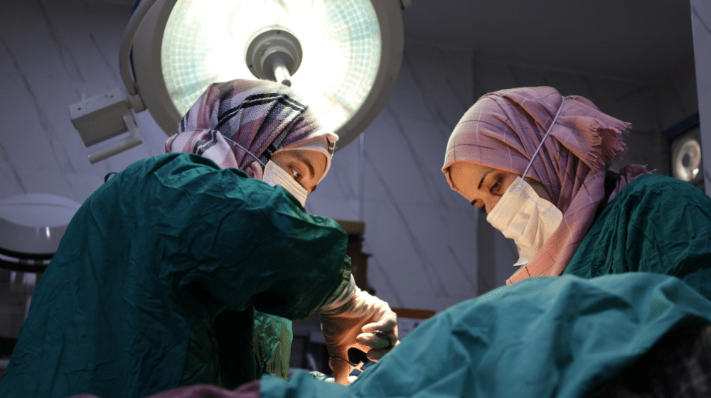 Two women medical workers wearing pink hijab and green operating gowns perform an operation under a large surgical light.