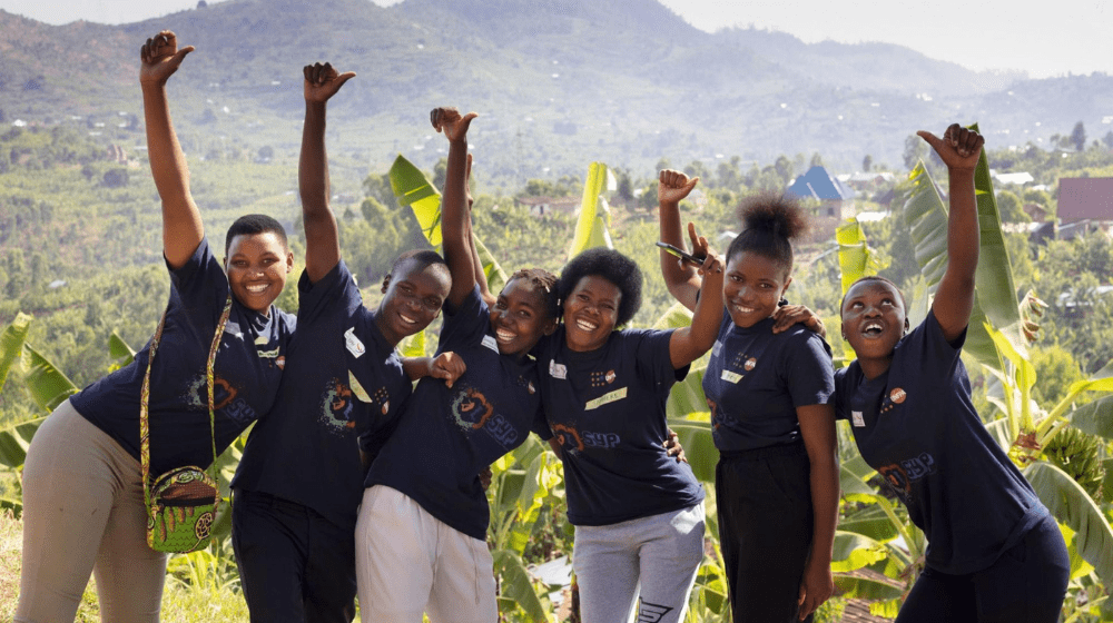 Six smiling young people all dressed in dark blue t-shirts raise their arms and embrace with greenery in the background.