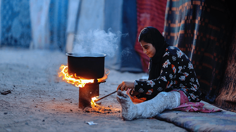 Teenage girl sits on the floor and kindles a fire for cooking. She has a broken leg. 