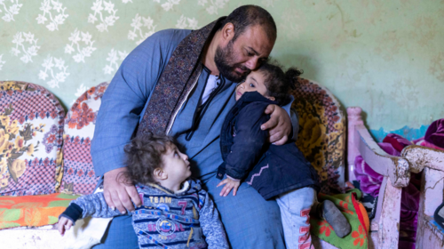 A man wearing a scarf and a blue tunic sits on a couch and embraces two young girls.