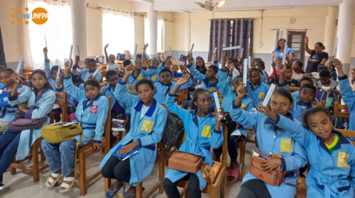 A group of young students in blue uniforms raise their hands while seated in a classroom