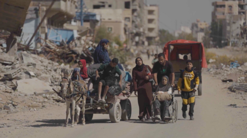 Un groupe de personnes, dont une en fauteuil roulant et plusieurs sur une carriole tirée par un âne, s’avancent sur une route poussiéreuse encadrée de bâtiments en ruines et de gravats